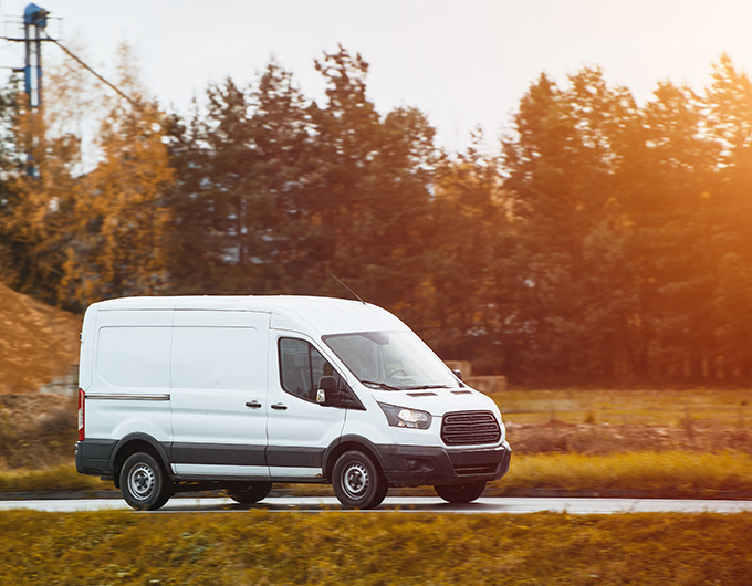 white cargo van driving on a road at sunset surrounded by trees with a modern design and spacious interior for transport of goods and supplies including 8-foot length options